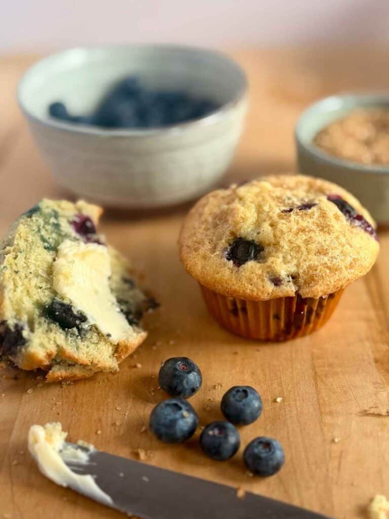 bakery style blueberry muffin cut in half with butter smeared on it and blueberries scattered around