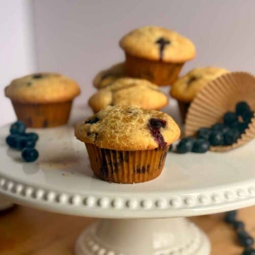 bakery-style blueberry muffins on white cake stand with blueberries in muffin liner in background