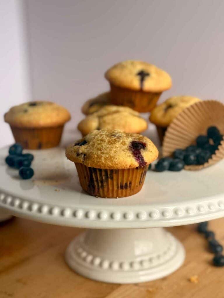 bakery-style blueberry muffins on white cake platter with blueberries falling out of a muffin liner in background 