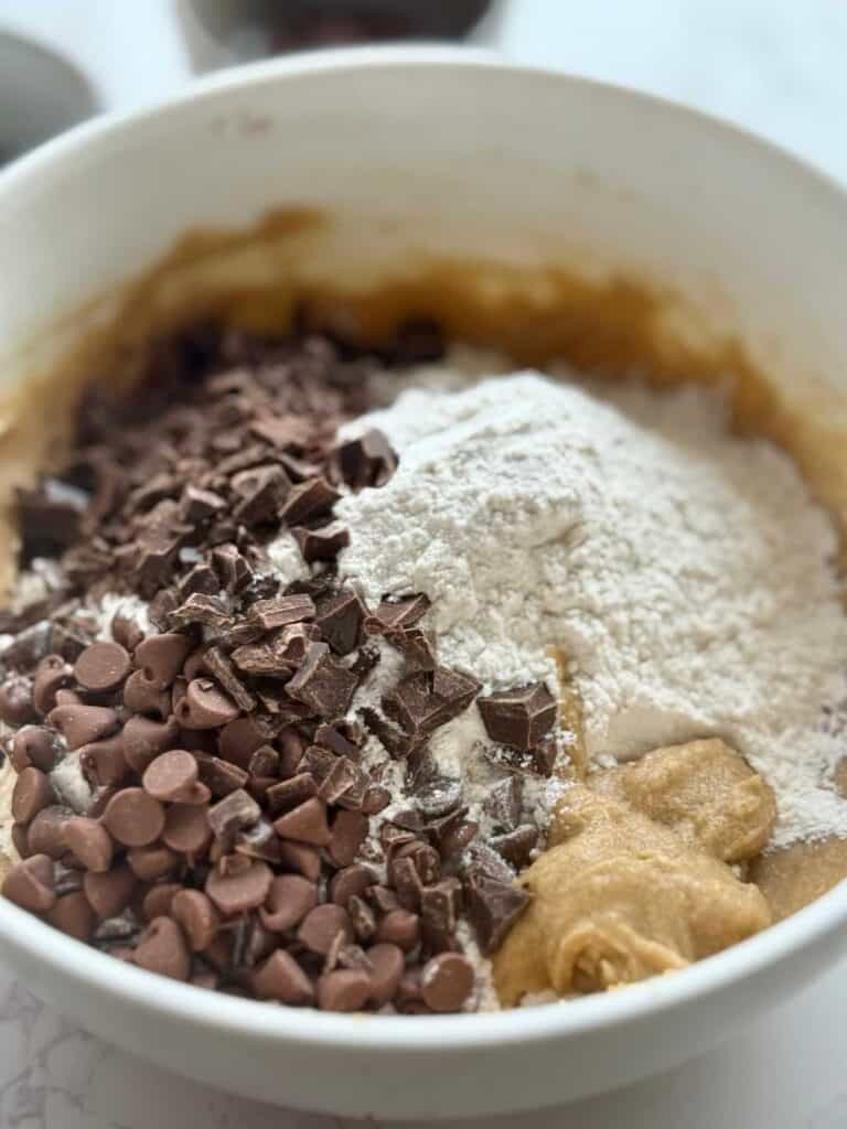 dry ingredients and chocolate chips and chunks in bowl on top of wet ingredients for sourdough brown butter chocolate chip cookies 