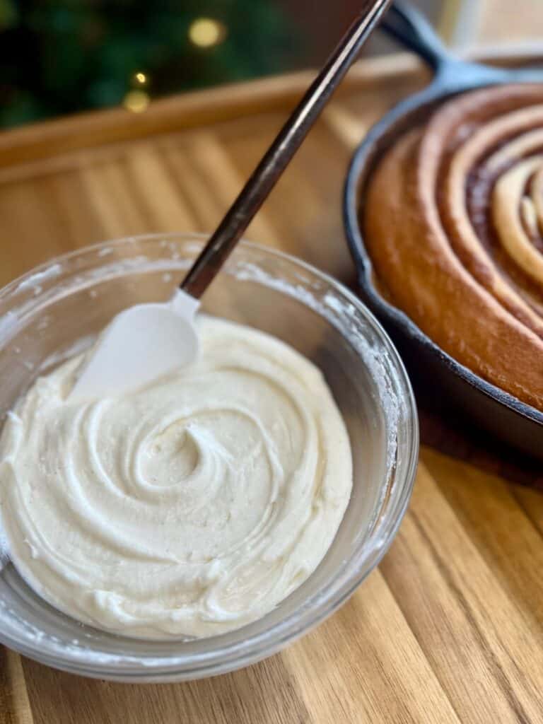 cast iron skillet cinnamon roll bowl of icing with spatula and cinnamon roll in background