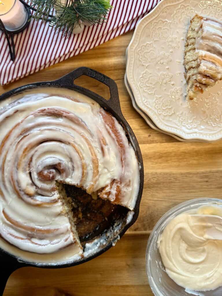 cast iron skillet cinnamon roll with slice cut out on plate Christmas deco and icing in background 
