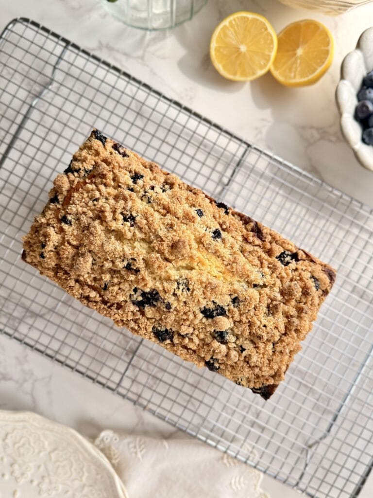 lemon blueberry loaf cake cooling on rack with fresh blueberries and lemons surrounding it