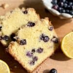 lemon blueberry loaf cake slices surrounded by fresh lemon slices and blueberries on wooden board