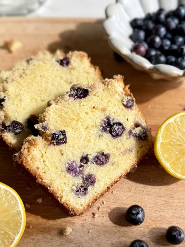 lemon blueberry loaf cake slices surrounded by fresh lemon slices and blueberries on wooden board