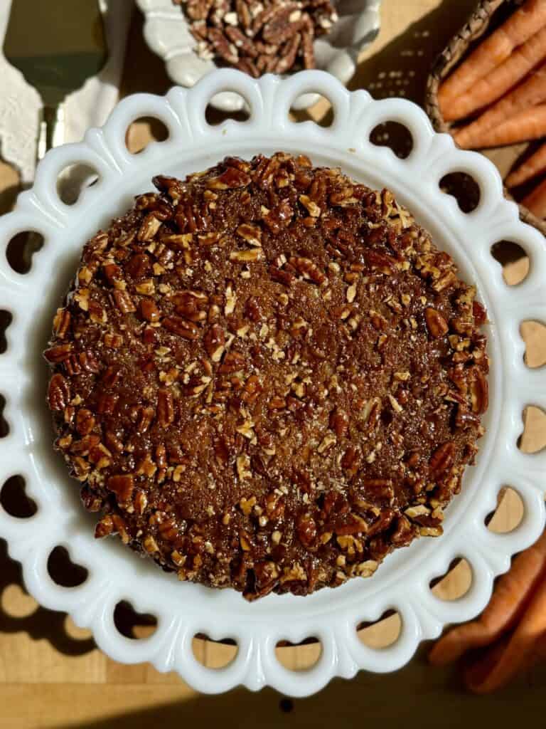 Sticky pecan carrot cake whole on serving plate with caramel pecan topping surrounded by carrots and pecans 
