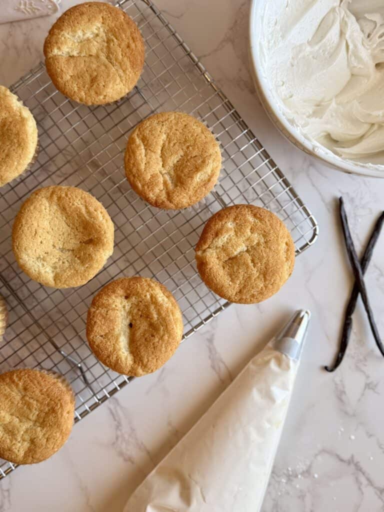 unfrosted vanilla cupcakes on cooling rack with buttercream in piping bag beside